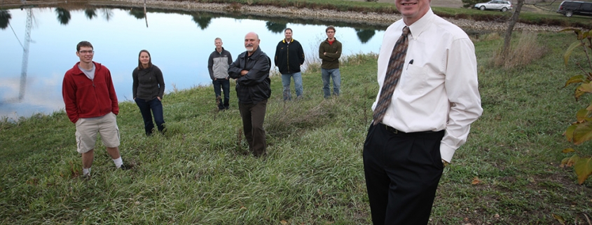 Larry Weber stands close to the camera with the rest of the IWP team behind him in front of a pond